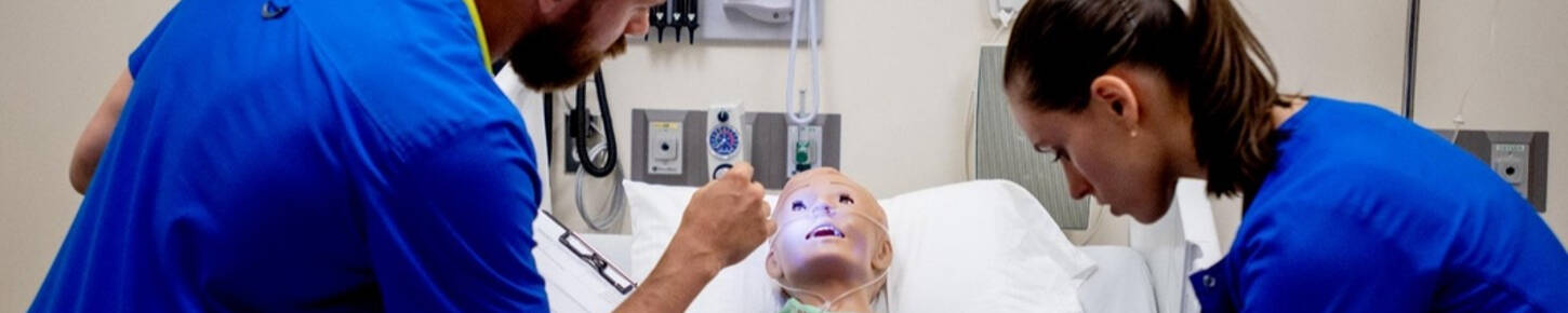 Two students in scrubs examine a manikin in a hospital bed